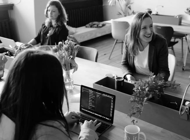 Two women working with laptops in an office while a third woman smiles and sits by their side looking off-camera.